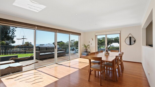 A spacious room with a large sliding glass door leading to an outdoor deck, a wooden dining table with chairs, and a round mirror on the wall. - Bodhi's Reef | Coastal Retreat (Lake Tyers Beach)