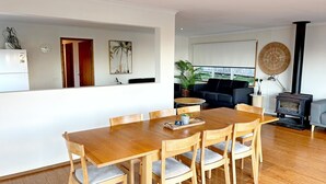 A dining area with a wooden table and chairs, a kitchen with a refrigerator, a living room with a sofa, and a wood-burning stove. - Bodhi's Reef | Coastal Retreat (Lake Tyers Beach)
