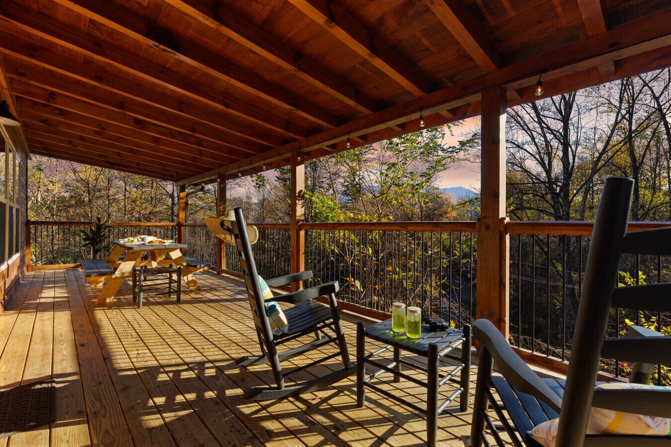 An outdoor wooden deck with tables and chairs, surrounded by a wooden railing, overlooking a forested area.