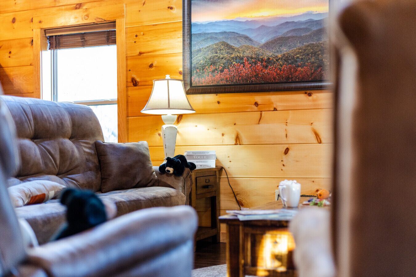 A cozy cabin interior with a plush sofa, a lamp, a painting of a mountainous landscape, and a small table with a cup and a teddy bear.