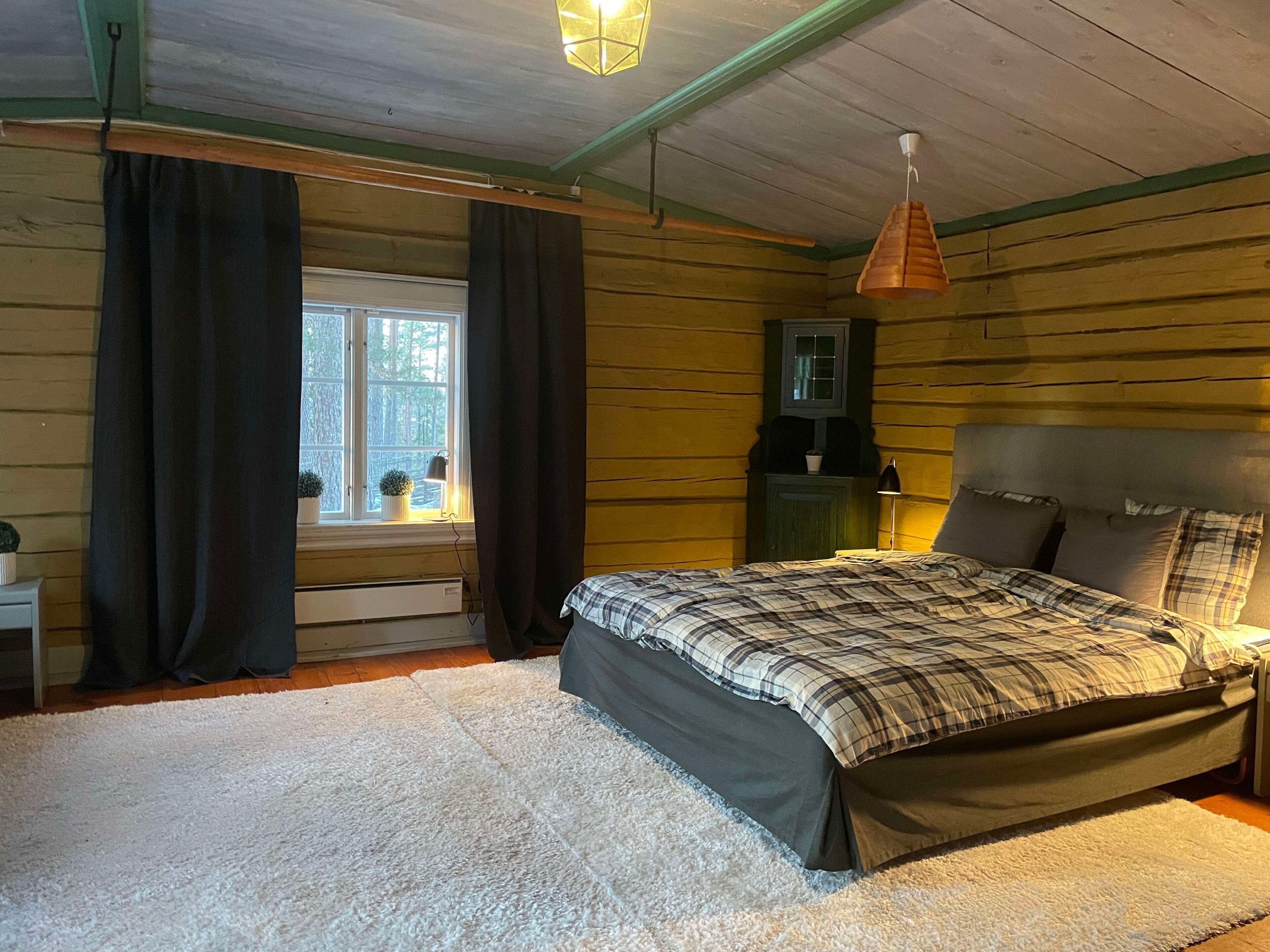 A bedroom with a bed, a window with curtains, a nightstand, a lamp, and a hanging light fixture.