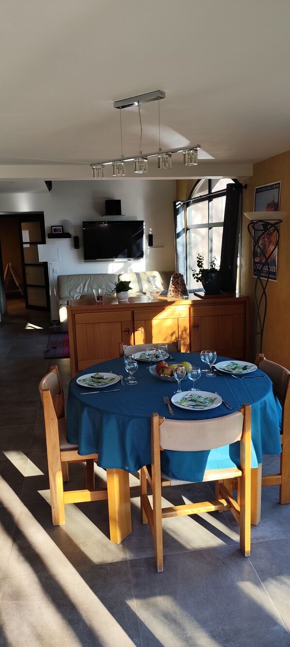 A dining area with a round table set for four, a television mounted on the wall, and a window with curtains.