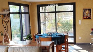 A modern living room with a sofa, dining table, and chairs. There are large windows allowing natural light and views of greenery outside.