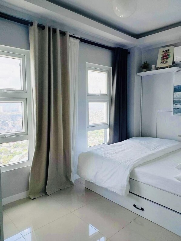 Un dormitorio con cama, dos ventanas, un armario y vistas a la ciudad.