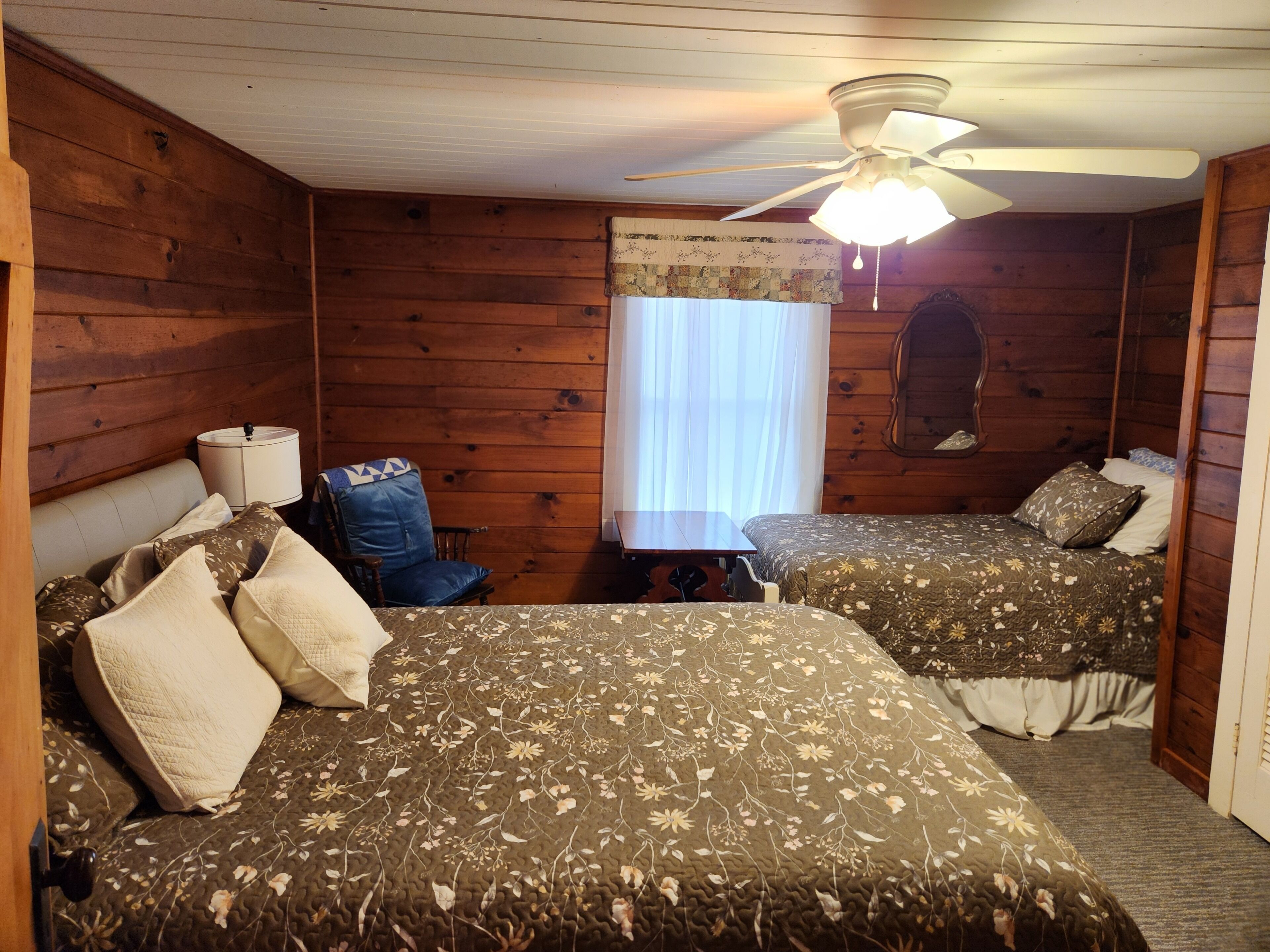 A room with two beds, a wooden ceiling, a ceiling fan, a table, and a chair.