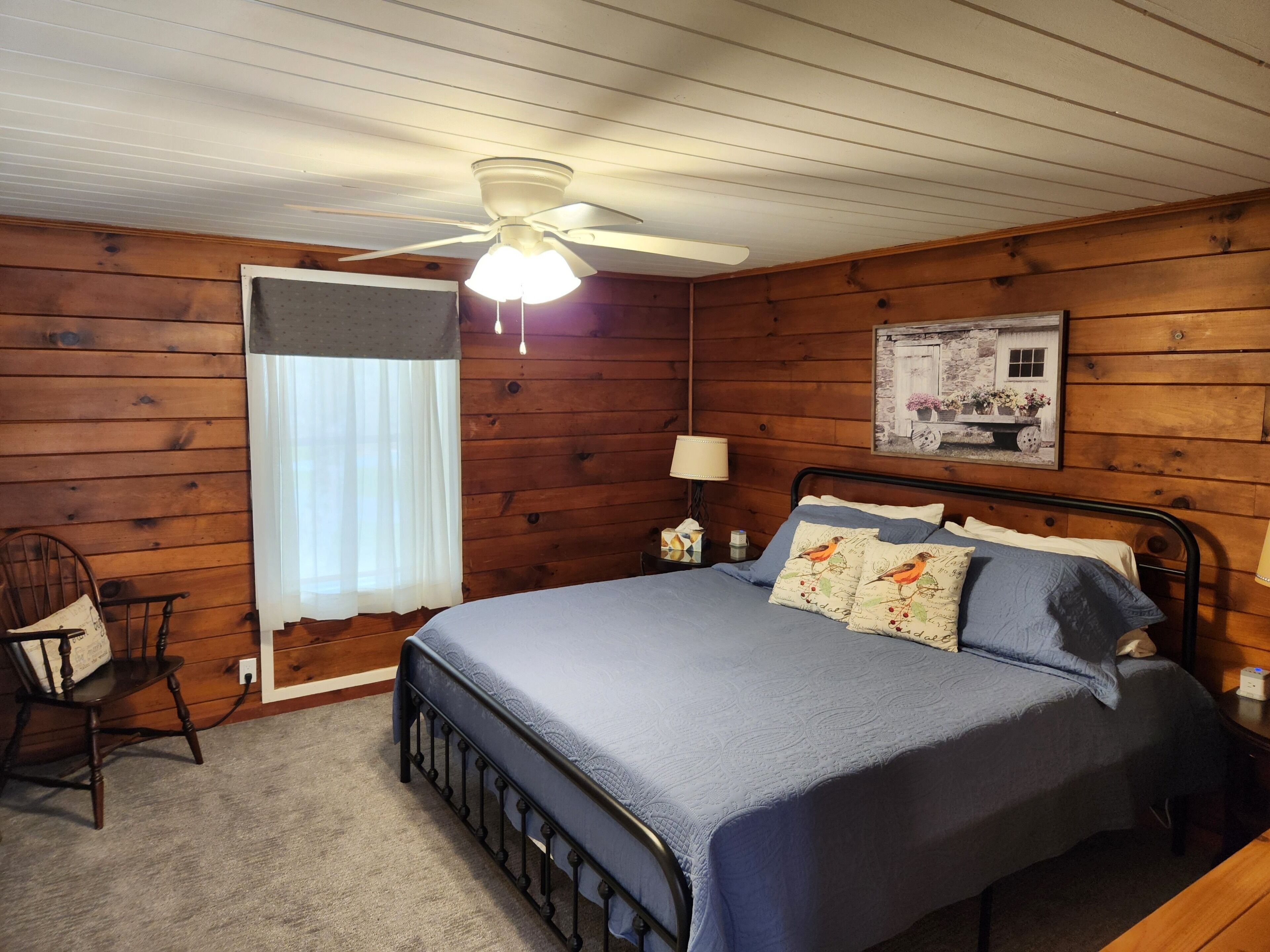 A wooden-paneled bedroom with a bed, a nightstand, a chair, a ceiling fan, and a window with curtains.