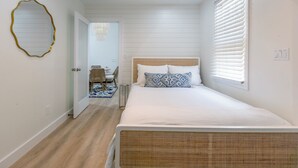 Une chambre avec un grand lit, un plancher en bois, un mur blanc et une fenêtre avec des stores.