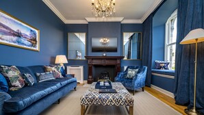 Un salon avec un canapé bleu, un pouf à motifs, un fauteuil bleu, une cheminée et un téléviseur fixé au mur.