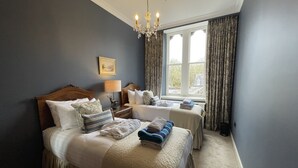 Une chambre d’hôtel avec un grand lit, une table de chevet avec une lampe, une fenêtre avec des rideaux et un lustre.
