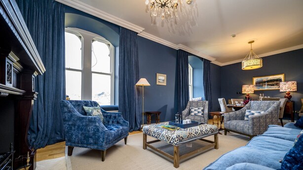 Un salon avec un canapé bleu, deux fauteuils, un pouf à motifs et un lustre.
