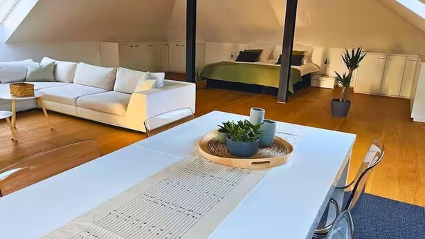 A modern loft bedroom with a sloped ceiling, a white sofa, a bed with a green bedspread, and a dining area with a table and chairs. - Modern loft in the center of Hattingen (Hattingen)