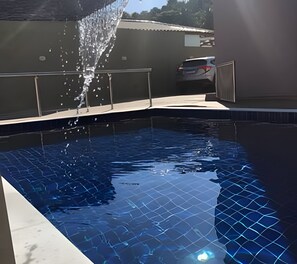 A swimming pool with clear blue water, a tiled deck, and a car parked in the background. - Condomínio com Piscina á 500m da Praia (Porto Seguro)