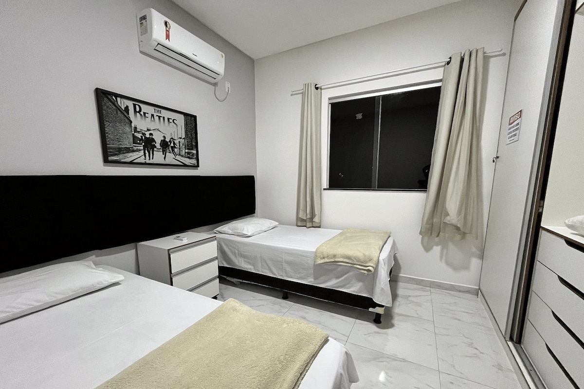 A hotel room with two single beds, a bedside table, a small desk, a window with curtains, and a wall-mounted air conditioner.