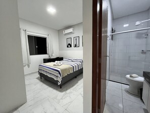 A modern bathroom with a shower, toilet, and a bed with a striped bedspread. - Condomínio com Piscina á 500m da Praia (Porto Seguro)
