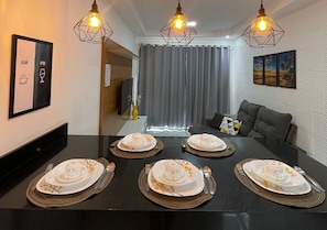 Un comedor con una mesa negra dispuesta para cuatro personas, tres lámparas colgantes, un cuadro enmarcado en la pared y una cortina gris al fondo.