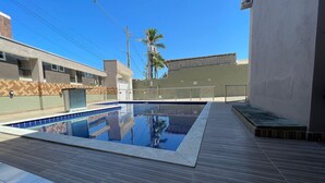 A swimming pool with a wooden deck, surrounded by buildings with a herringbone pattern wall and a palm tree. - Condomínio com Piscina á 500m da Praia (Porto Seguro)