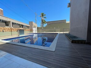A swimming pool with a wooden deck, surrounded by buildings with a herringbone pattern wall and a palm tree. - Condomínio com Piscina á 500m da Praia (Porto Seguro)