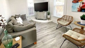 Een moderne woonkamer met een grijze bank, rieten stoelen, een flatscreen televisie en een salontafel.