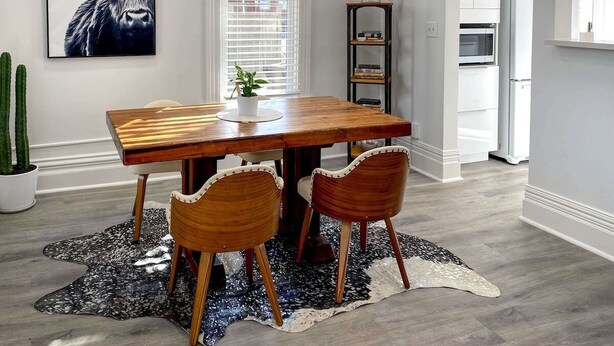 Een eetruimte met een houten tafel en twee stoelen, een potplant, een zwart-wit vloerkleed, en een kunstwerk met stieren.