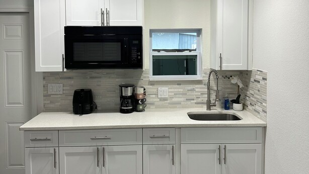A modern kitchen with white cabinets, a black microwave, a coffee maker, and a window above the sink. - Charming Studio Apartment at Lake Worth Beach! (Lake Worth Beach)