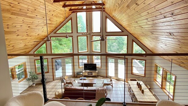 A spacious loft with a high ceiling, large windows, and a cozy seating area with a TV. - Modern A-Frame Cabin, Sleeps 12, Cedar Hot Tub, Game Room, Fishing Pond, Views (Ellijay)