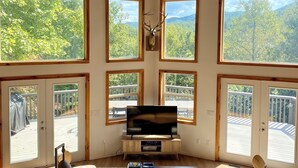 A spacious living room with large windows, a chandelier, and a mounted deer head. - Modern A-Frame Cabin, Sleeps 12, Cedar Hot Tub, Game Room, Fishing Pond, Views (Ellijay)