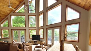 A spacious living area with a high ceiling, large windows, a wooden dining table, and a cozy seating arrangement. - Modern A-Frame Cabin, Sleeps 12, Cedar Hot Tub, Game Room, Fishing Pond, Views (Ellijay)