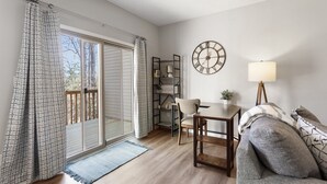 Ein gemütliches Wohnzimmer mit Essbereich, einem großen Fenster mit Vorhängen, einer Uhr an der Wand und einer Lampe.