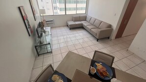 Una sala de estar moderna con un sofá seccional, una mesa de comedor con desayuno, y un ventanal grande.