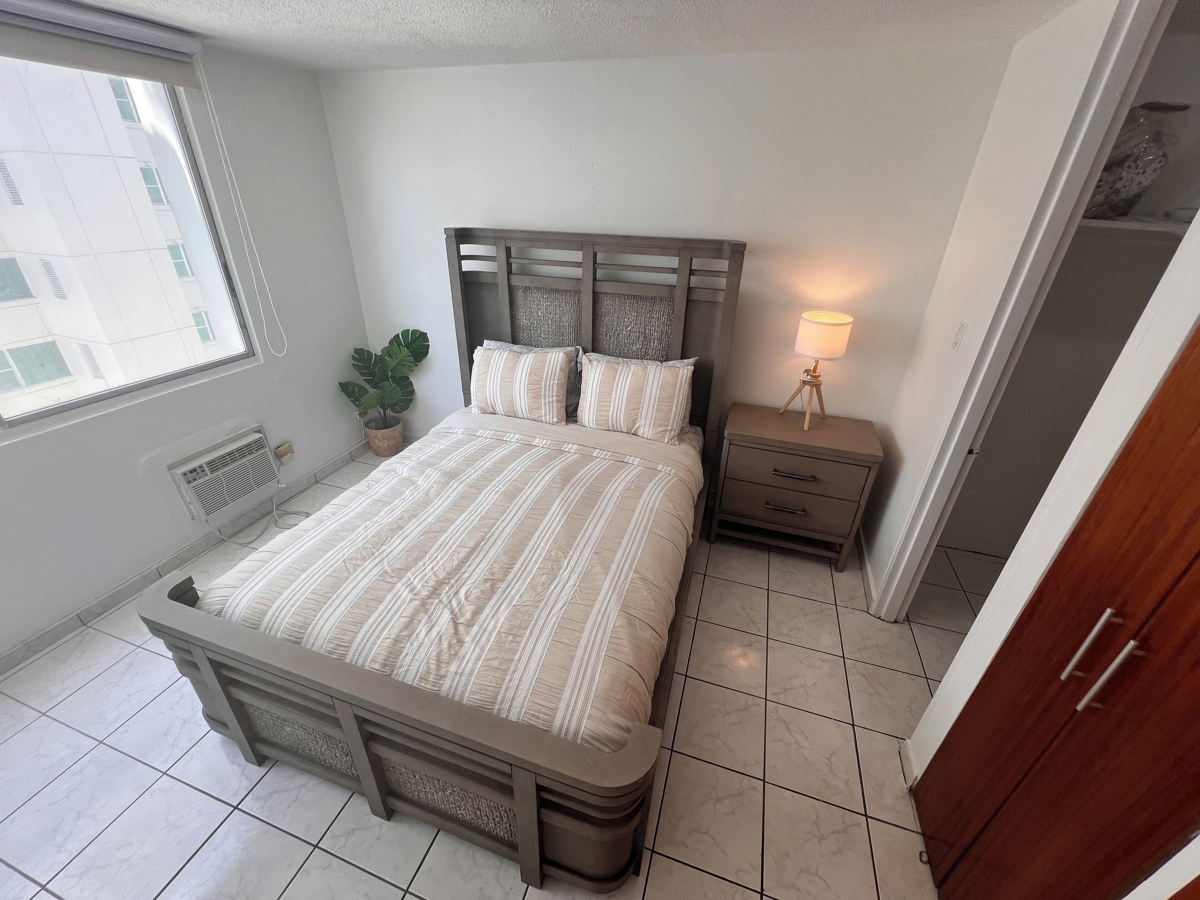 A bedroom with a bed, bedside table, lamp, potted plant, and a view of buildings outside.