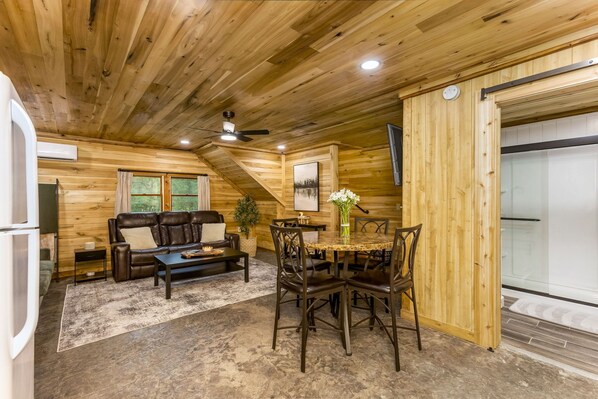 A cozy cabin interior with a wooden ceiling, a dining area with a round table and chairs, a leather sofa, and a small side table. - Creekside Cabin in Hocking Hills. Hot Tub + Large Private Yard. Dog-friendly! (Rockbridge)