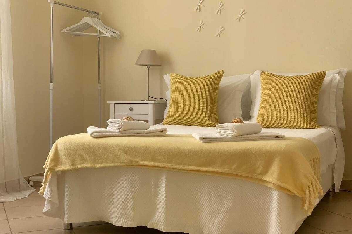 Una cama bien tendida con ropa de cama blanca y detalles amarillos, una mesita de noche con lámpara, y una lámpara de pie.