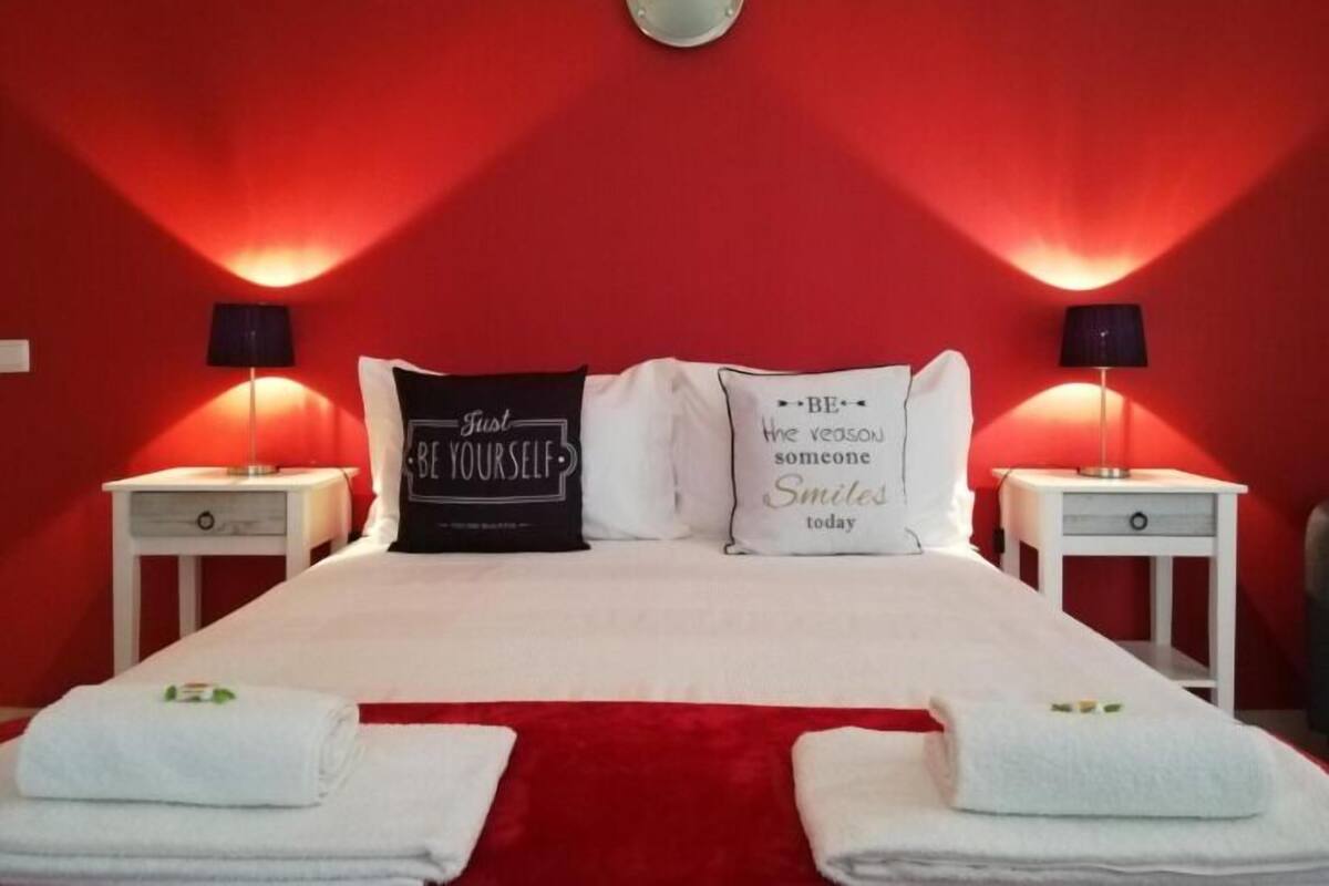 Habitación de hotel con pared roja, dos mesitas de noche con lámparas, cama con ropa blanca y dos almohadas con citas motivacionales.
