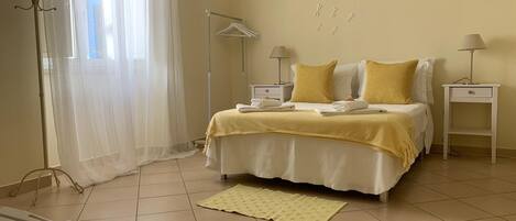 A bedroom with a bed, bedside tables, a lamp, a hanging coat rack, and a window with sheer curtains.