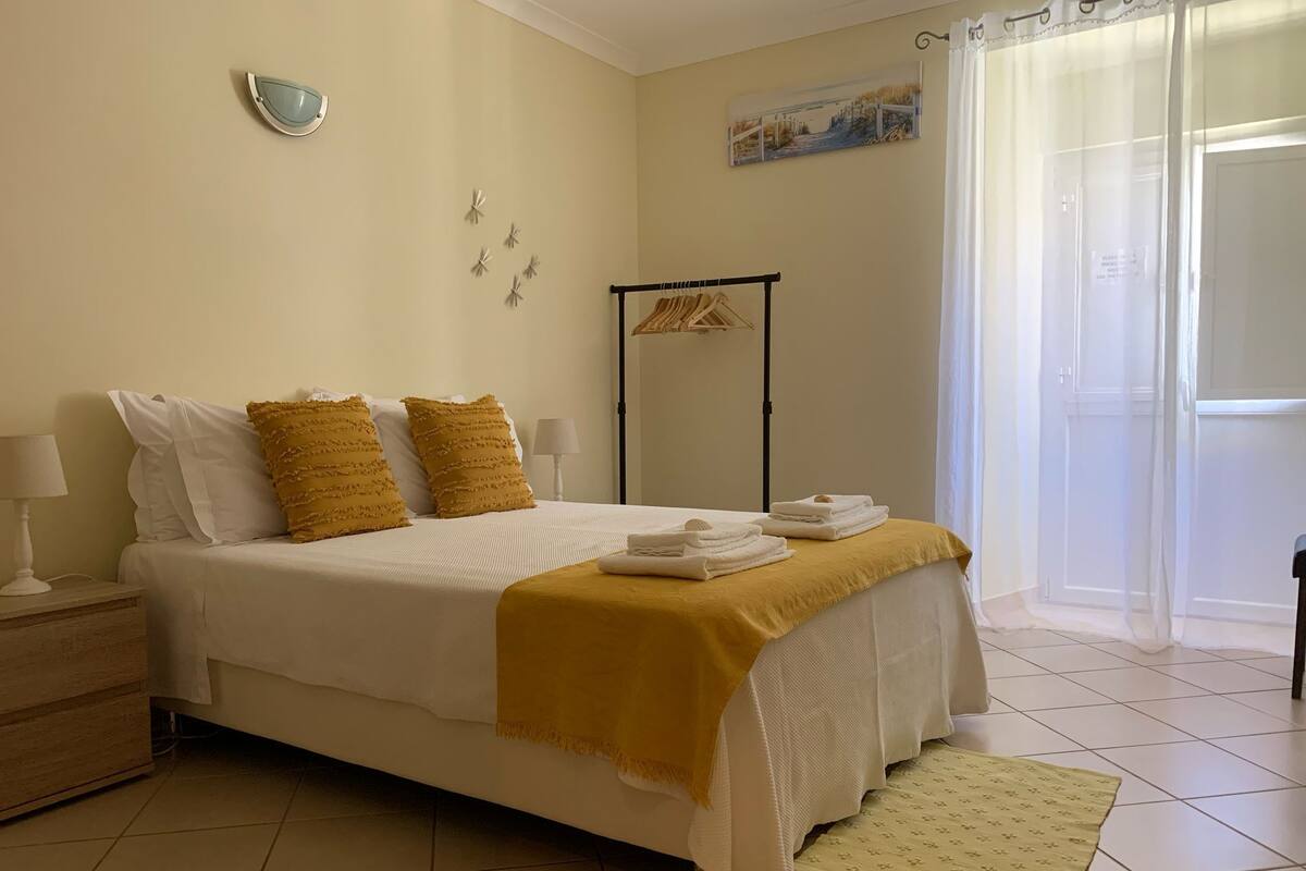 Un dormitorio ordenado con una cama, almohadas, una mesita de noche, una lámpara, un perchero para toallas y un cuadro en la pared.