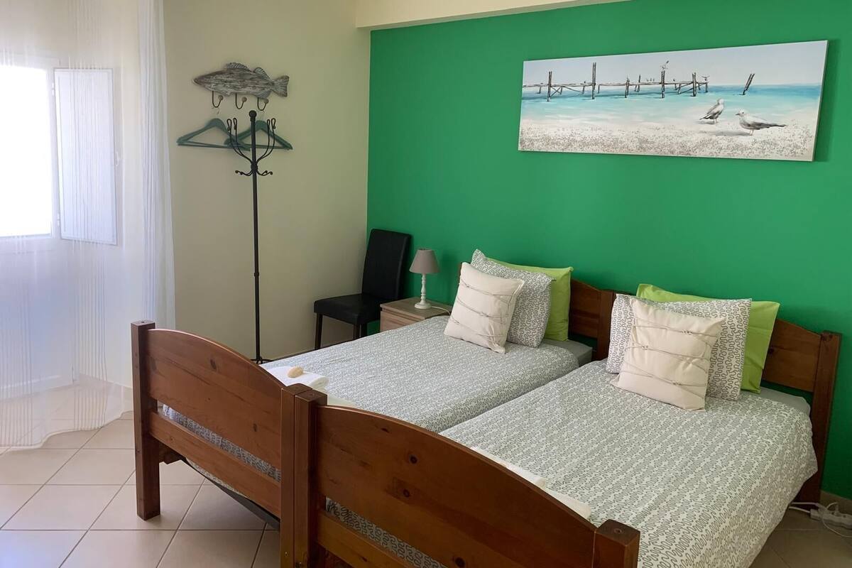 Un dormitorio con dos camas, una pared verde, un cuadro de una playa y un perchero con detalle de pez.