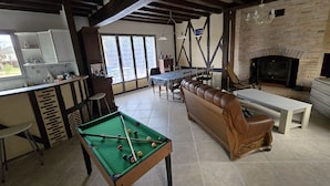 Een ruime woonkamer met een pooltafel, een bank, een eettafel met stoelen, een open haard en een keukenhoek.
