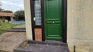 Een groene deur met een bakstenen basis, een klein raam met bloemenmotief, en een betonnen pad dat naar een gebouw met pannendak leidt.