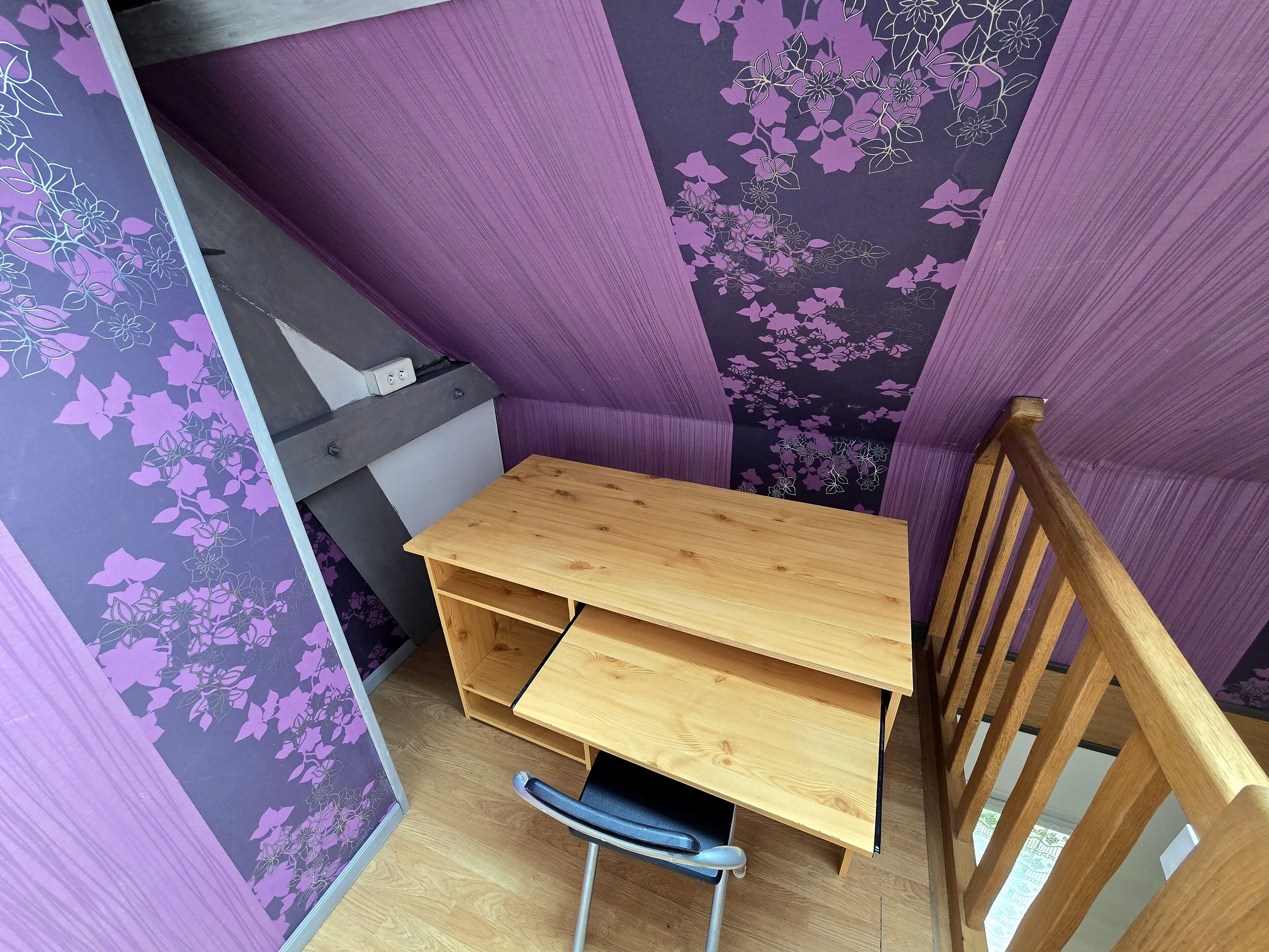 A corner room with a wooden desk, a chair, and purple floral wallpaper.