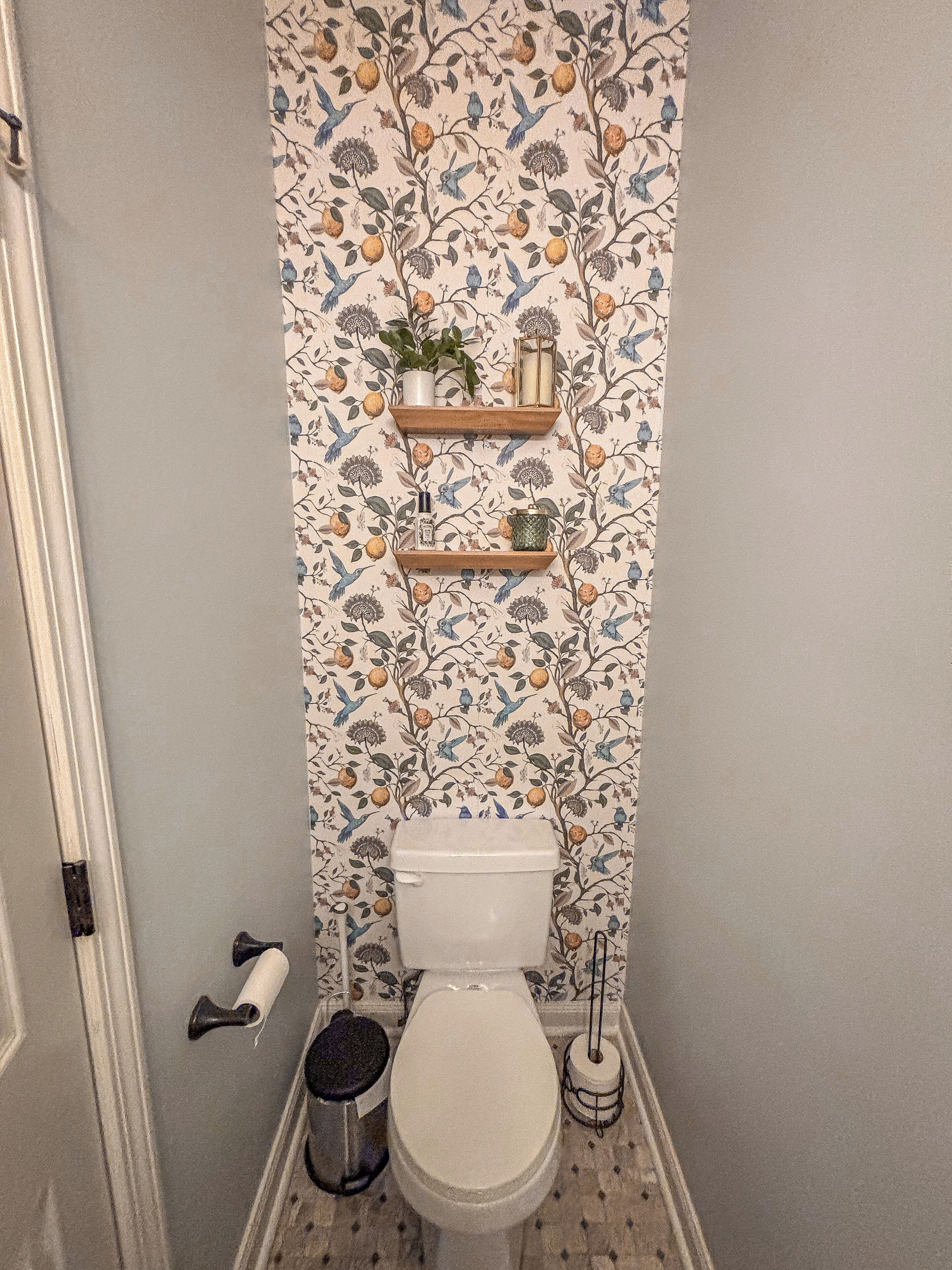 A bathroom with a floral wallpaper, two wooden shelves with decorative items, a toilet, and a trash bin.