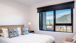 A bedroom with a large window, a bed with patterned pillows, a bedside table with a lamp, and a chair. - Drop In Condo by Revelstoke Vacations (Revelstoke)