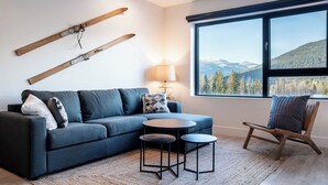 A modern living room with a sectional sofa, two small tables, a wooden chair, and a large window with a mountain view. - Drop In Condo by Revelstoke Vacations (Revelstoke)
