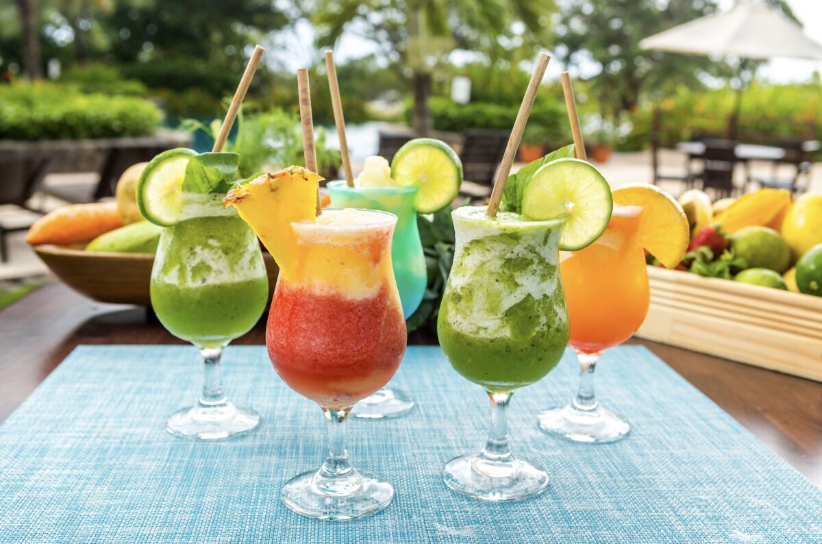 Trois cocktails colorés, décorés de quartiers de citron vert, sont disposés sur une table, entourés de fruits et d’un saladier rempli d’autres fruits.