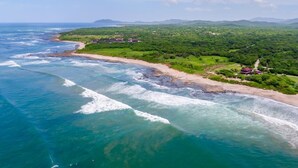 Vista aérea de una zona costera con playa, vegetación y algunos edificios.