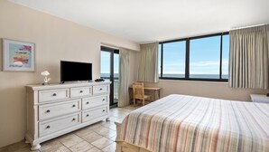 Een hotelkamer met een bed, een televisie op een dressoir, een stoel en uitzicht op zee door grote ramen.