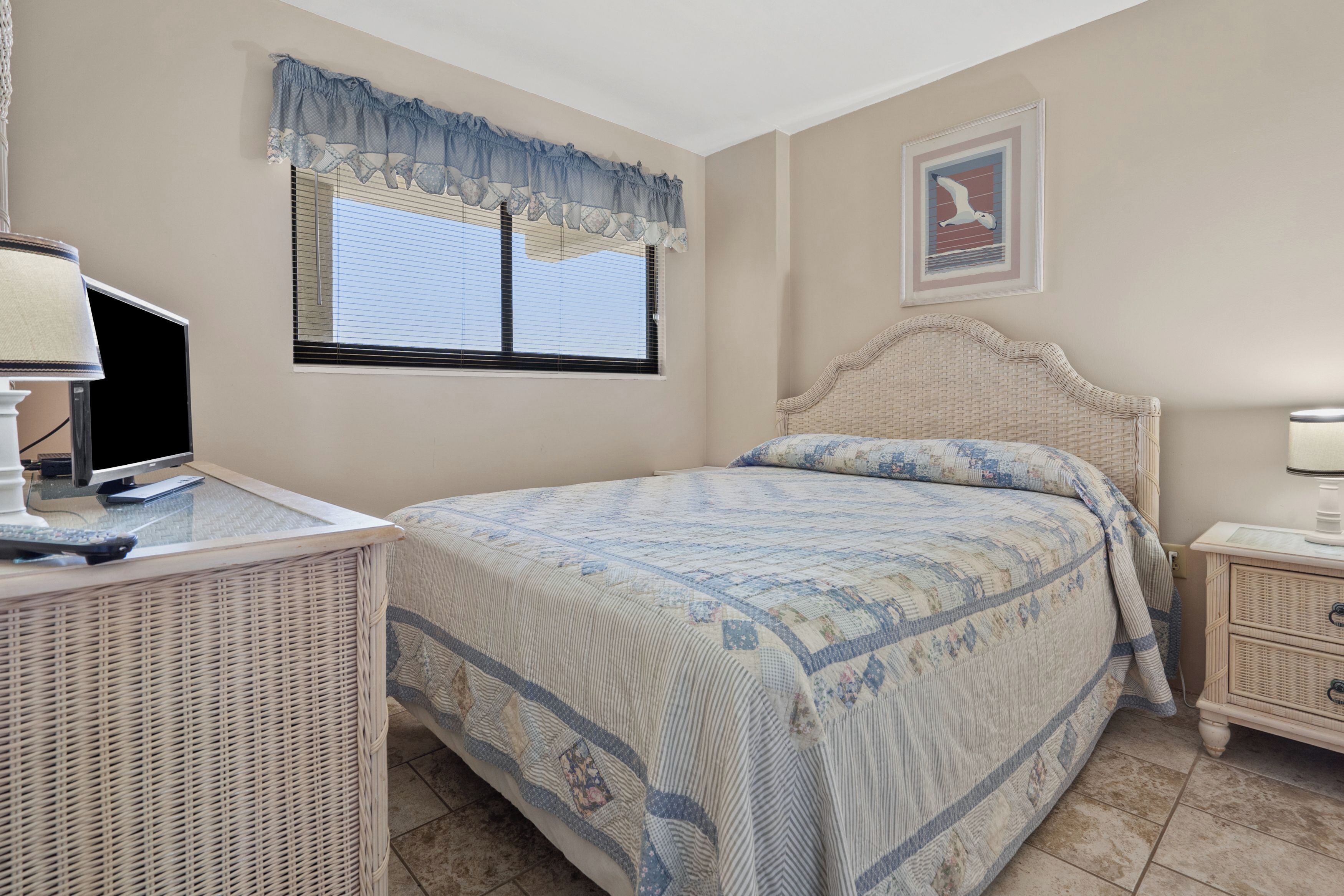 Un dormitorio con cama, escritorio con computadora, silla de mimbre, mesita de noche y ventana con persianas.