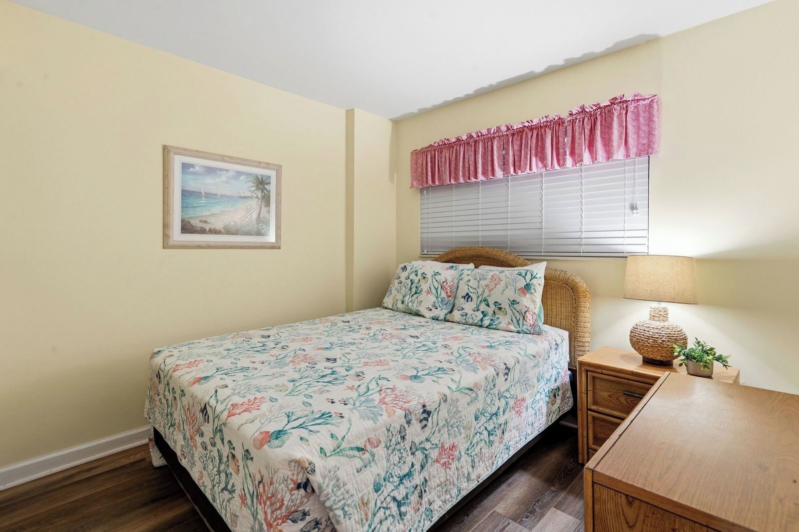 A bedroom with a bed, a bedside table with a lamp, a window with blinds, and a framed picture on the wall.