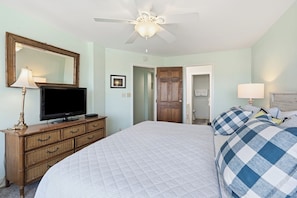 A bedroom with a bed, a television on a dresser, a ceiling fan, and a door leading to a bathroom. - Sedgefield 102 (North Myrtle Beach)