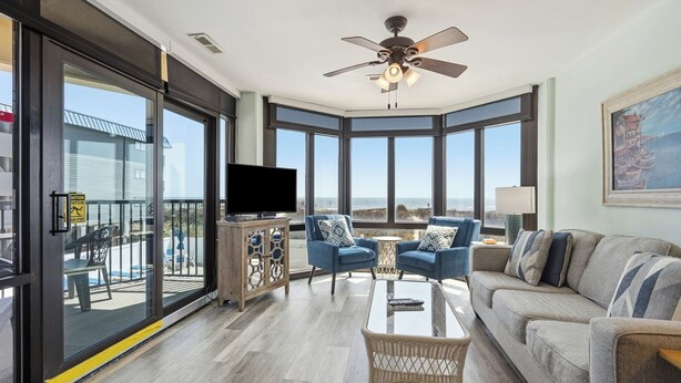 A spacious living room with a large TV, comfortable seating, a ceiling fan, and a balcony with a view. - Sedgefield 102 (North Myrtle Beach)