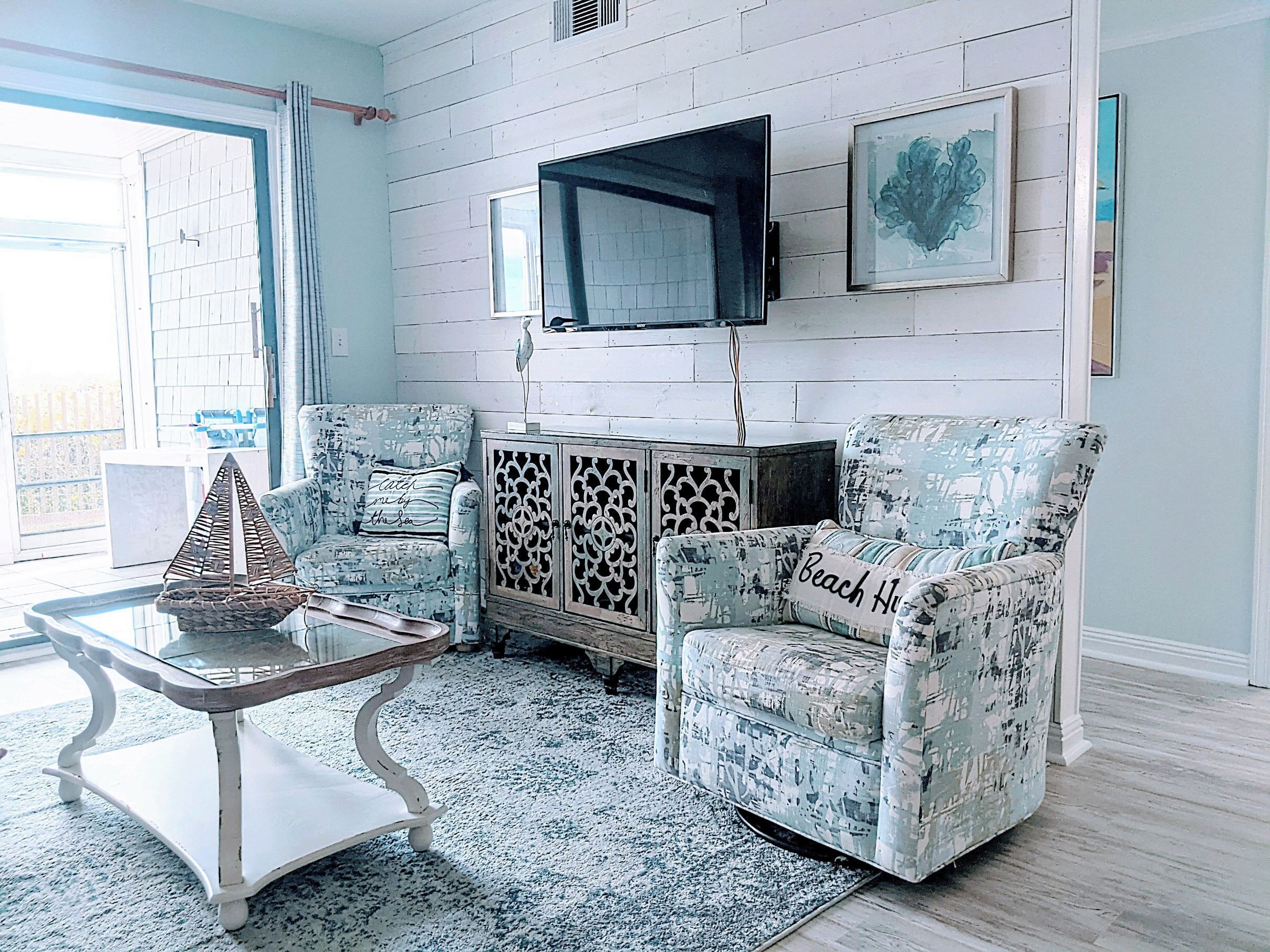 A living room with two patterned armchairs, a glass coffee table, a wooden cabinet, and a mounted TV.